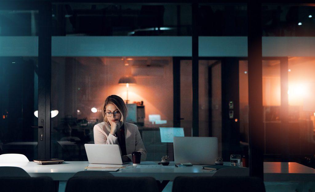 Businesswoman working late at night in a modern office, focused on her laptop with a serious expression.